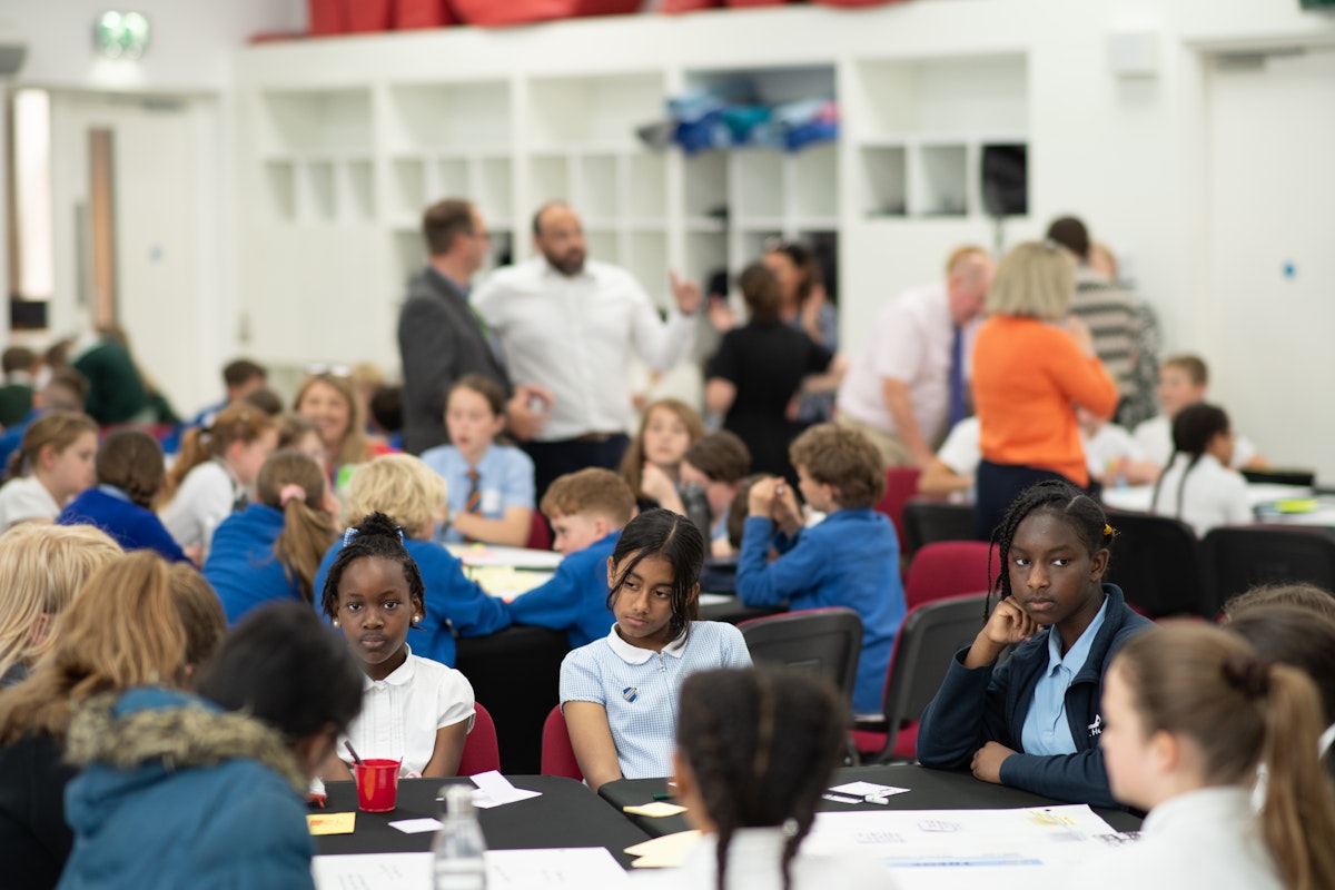 Children in school uniform sit in groups around large tables, focusing on written ideas and discussions during the Hope Hack